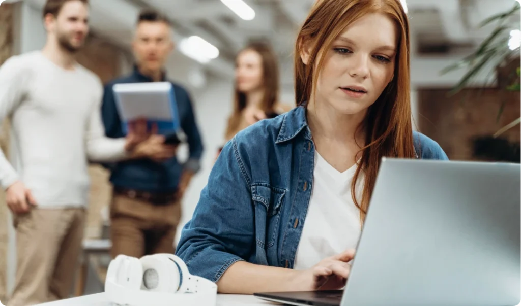 Young person with long red hair working on a laptop in a modern office; white headphones on the table, with several people talking in the background